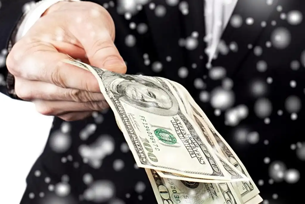 A person in a suit extends their hand forward, holding several U.S. hundred-dollar bills, with white circular light spots in the background, giving a festive or celebratory atmosphere.