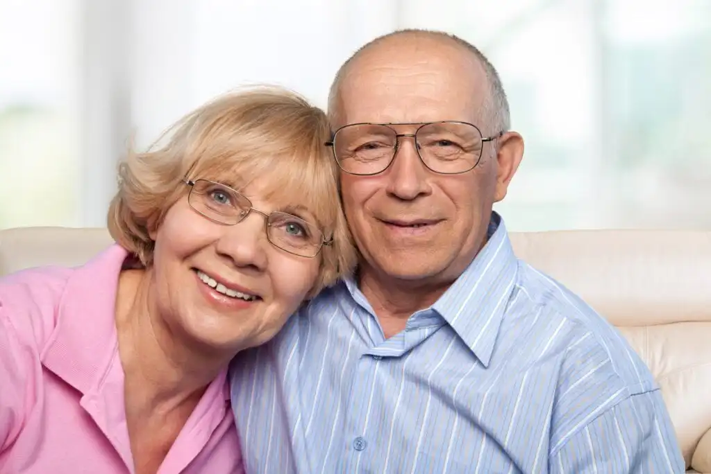 An elderly couple sits close together on a couch, smiling warmly at the camera. The woman wears glasses and a pink shirt, and the man wears glasses and a light blue striped shirt. The background is softly blurred.