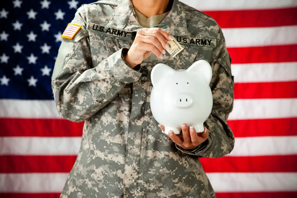 A person in a U.S. Army uniform holds a white piggy bank and puts a dollar bill into it, with the American flag in the background.