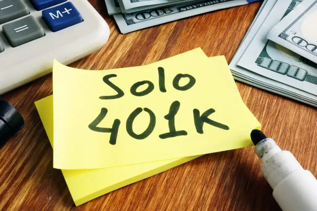 A yellow sticky note with Solo 401k written on it is on a wooden desk, next to a marker, a calculator, and stacks of cash.