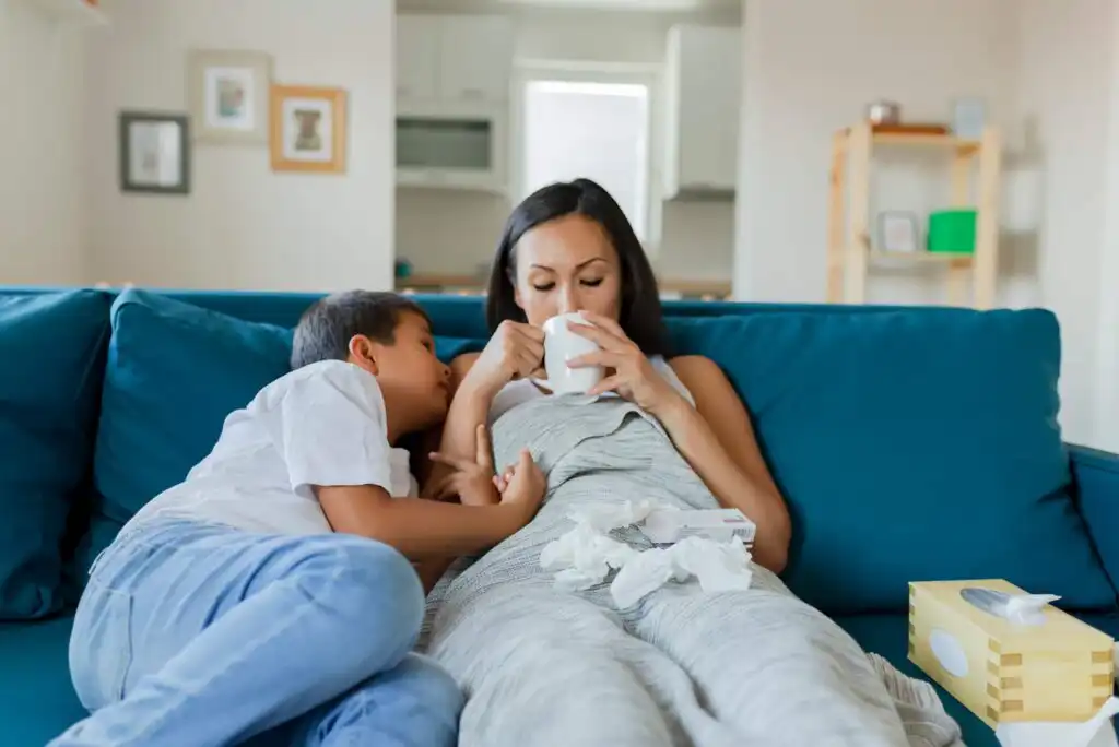 A woman wrapped in a blanket sits on a blue couch drinking from a mug, with a young boy leaning against her. Used tissues and a tissue box are on the couch, suggesting she is unwell.