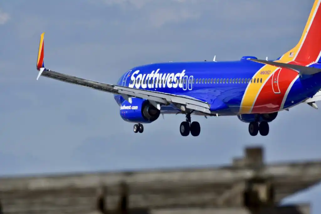 A Southwest Airlines jet with blue, red, and yellow colors is landing with its landing gear down, flying low over a blurred structure in the foreground against a cloudy sky.