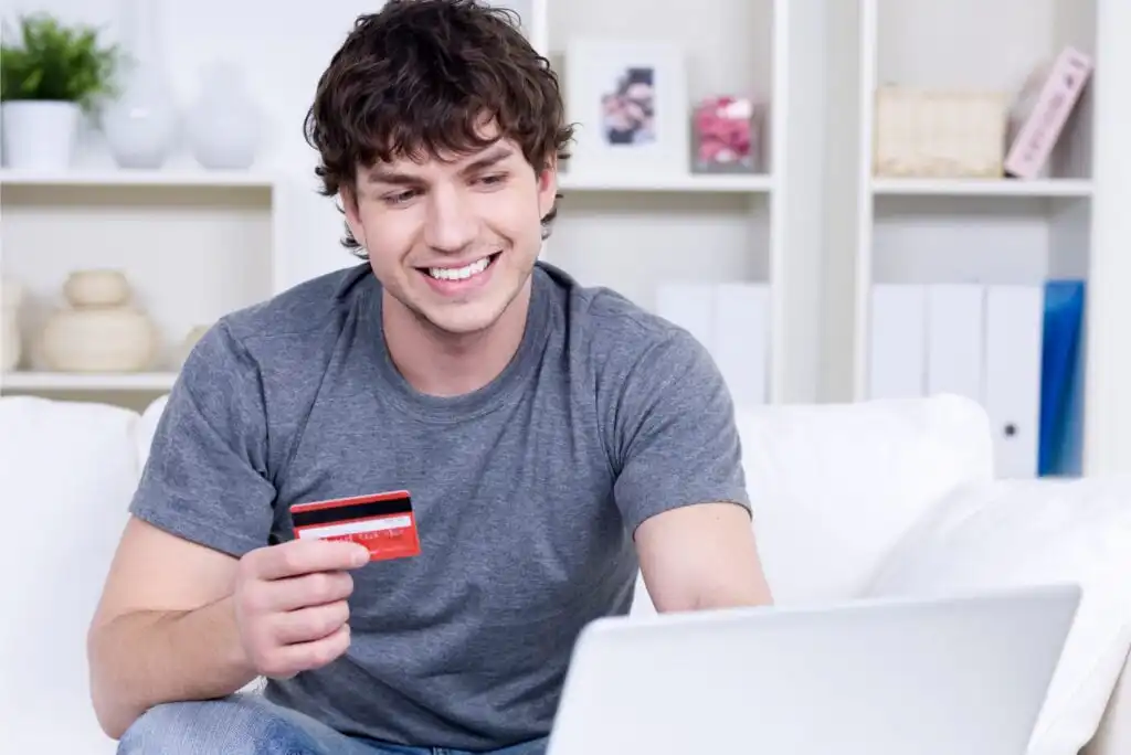 A smiling young man in a gray t-shirt sits on a couch holding a red credit card in one hand and looks at a laptop in front of him, suggesting he is shopping or banking online at home.