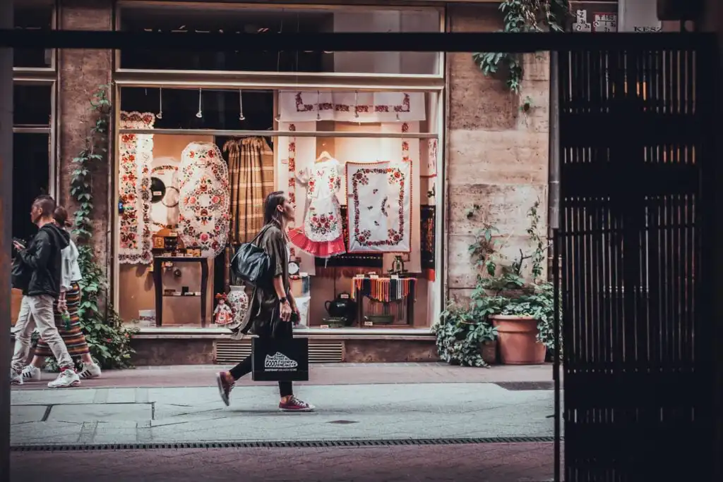 A person walks past a shop window displaying colorful textiles and clothing; they carry a shopping bag and wear a backpack. The street is lined with plants and the shop’s interior is warmly lit.