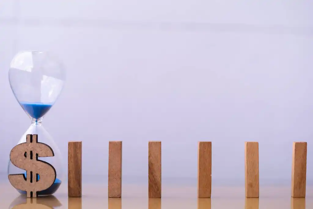 A glass hourglass filled with blue sand stands next to a wooden dollar sign and five upright wooden domino pieces, all arranged in a row on a reflective surface against a light background.