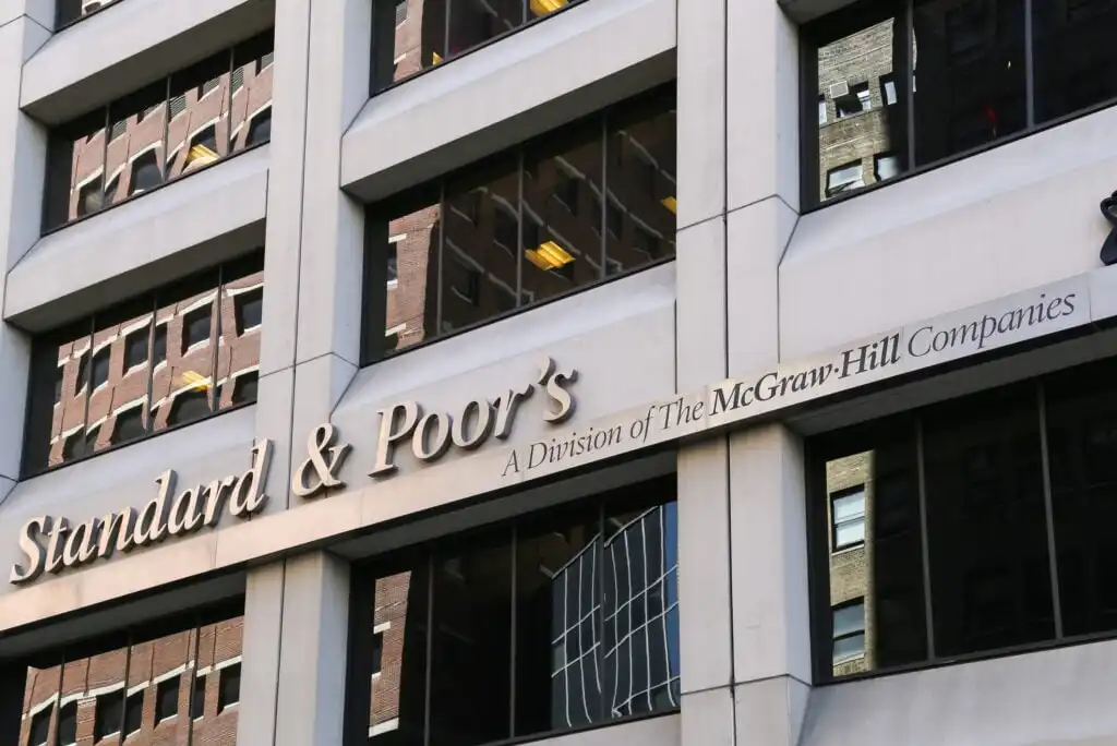 The exterior of a building displays the sign “Standard & Poor’s, A Division of The McGraw-Hill Companies,” with reflections of other buildings visible in the windows.