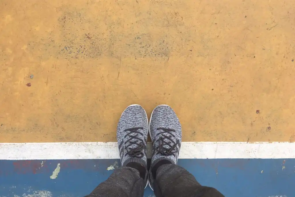 Person wearing gray sneakers and dark pants standing on a white line that separates a blue area from a yellowish concrete surface, viewed from above.