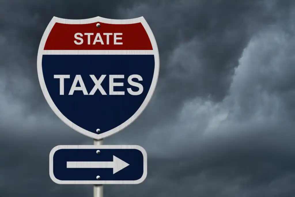 A road sign with STATE TAXES written on it stands against a backdrop of dark, cloudy skies, with an arrow pointing to the right below the text.