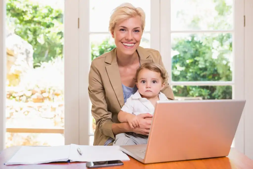A smiling woman sits at a desk with a laptop, holding a young child on her lap. Papers, a pen, and a smartphone are on the desk. Bright windows and greenery are visible in the background.