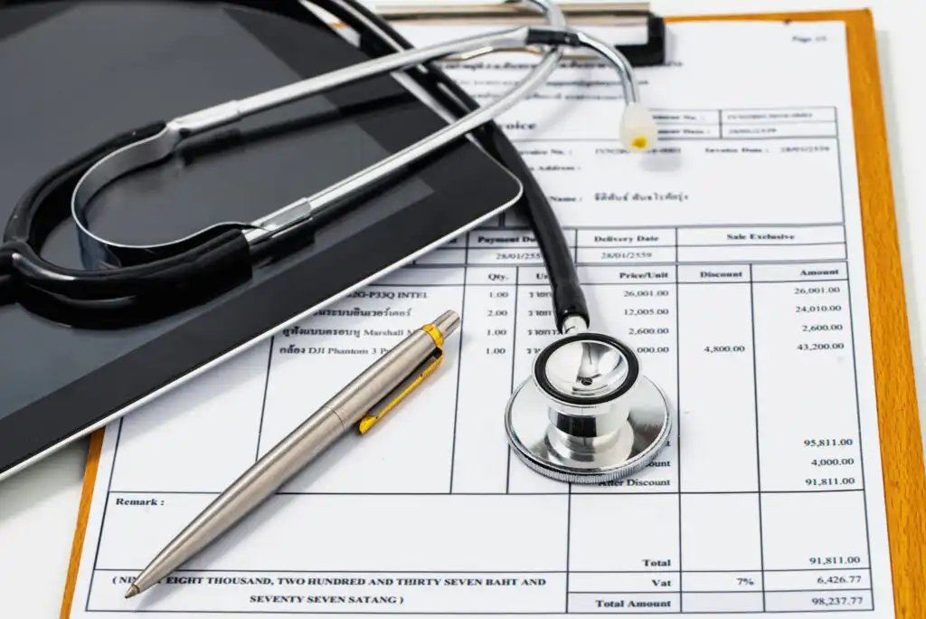 A stethoscope, pen, and clipboard with medical documents and a tablet are arranged on a white surface, symbolizing healthcare, medical billing, or health insurance paperwork.
