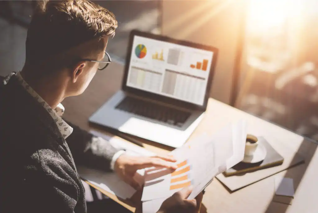 A man wearing glasses analyzes charts and graphs on paper and a laptop at a desk with sunlight streaming in. A cup of coffee and notebook are also on the desk.
