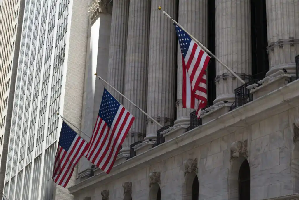 Three American flags at half-mast hang in front of a large stone building with tall columns and arched windows, likely a government or financial institution, in an urban setting.