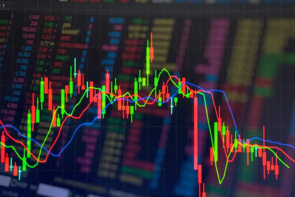 Colorful stock market candlestick chart with green, red, and blue lines indicating price changes, displayed against a blurred background of numbers and financial data on a digital screen.