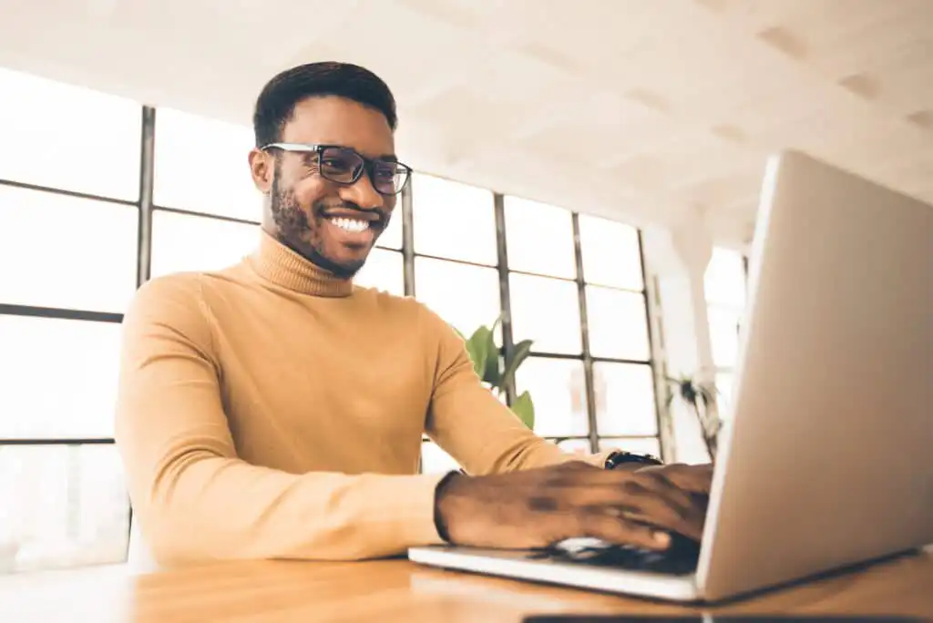 A smiling man wearing glasses and a beige turtleneck sweater sits at a desk, typing on a laptop in a bright, modern office with large windows in the background.