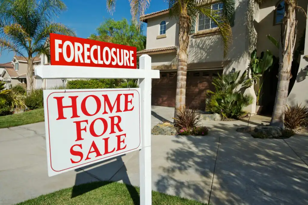 A “Home for Sale” sign, with a red “Foreclosure” notice above it, stands in front of a beige suburban house with a brown garage door, palm trees, and landscaped front yard.