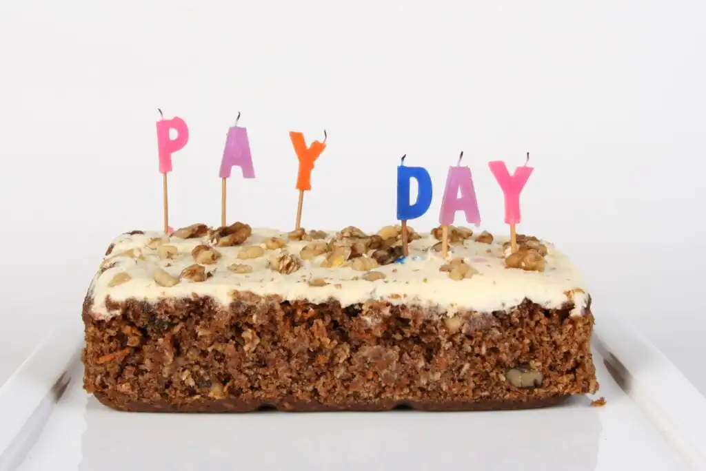 A rectangular cake with white icing and walnuts on top, decorated with colorful letter candles spelling PAY DAY against a plain white background.