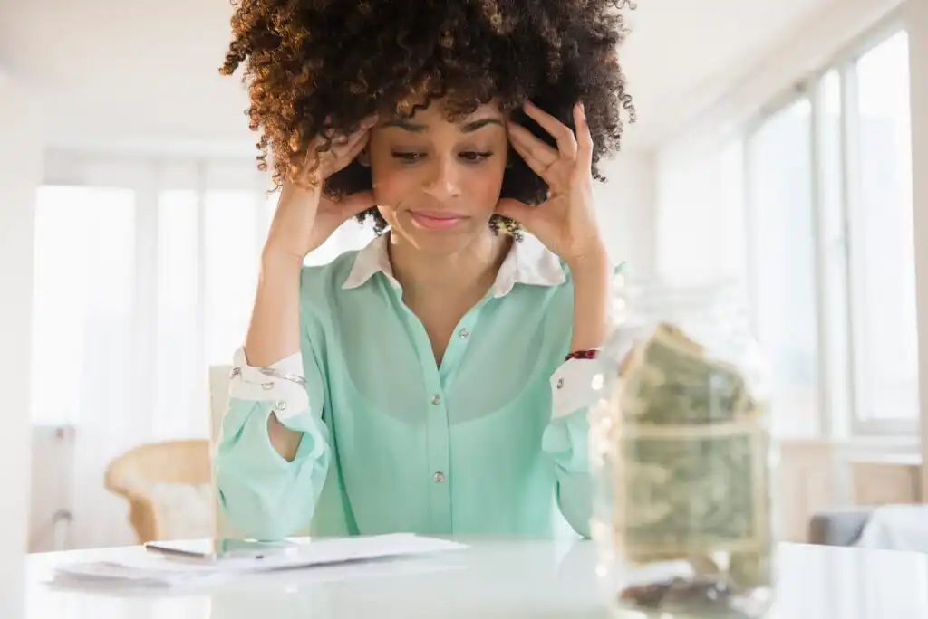 A woman sits at a table looking stressed, with her hands on her temples. In front of her is a glass jar filled with cash and some papers, suggesting financial concern. Bright daylight fills the room.