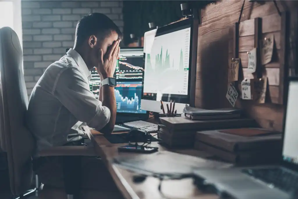 A man sits at a cluttered desk with multiple monitors displaying financial charts, holding his head in his hands, appearing stressed or frustrated in an office setting.