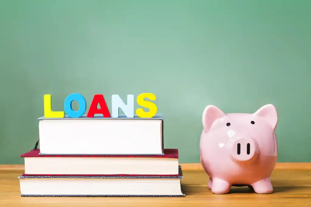 A pink piggy bank sits next to a stack of books with colorful letters on top spelling LOANS, all set against a green background.