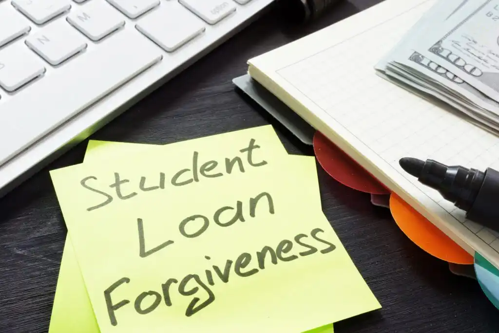 A yellow sticky note with Student Loan Forgiveness written on it sits on a dark desk near a keyboard, a notebook, a black marker, and a partial view of cash.