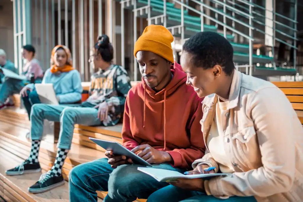 Two students sit on a bench indoors, working together with a tablet and notebook. Other students are seated in the background, some using laptops, near a staircase in a modern, open study space.