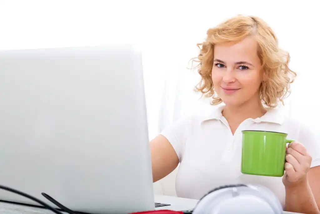 A woman with curly blonde hair sits at a desk using a laptop and holds a green mug, smiling at the camera. The background is bright and white.