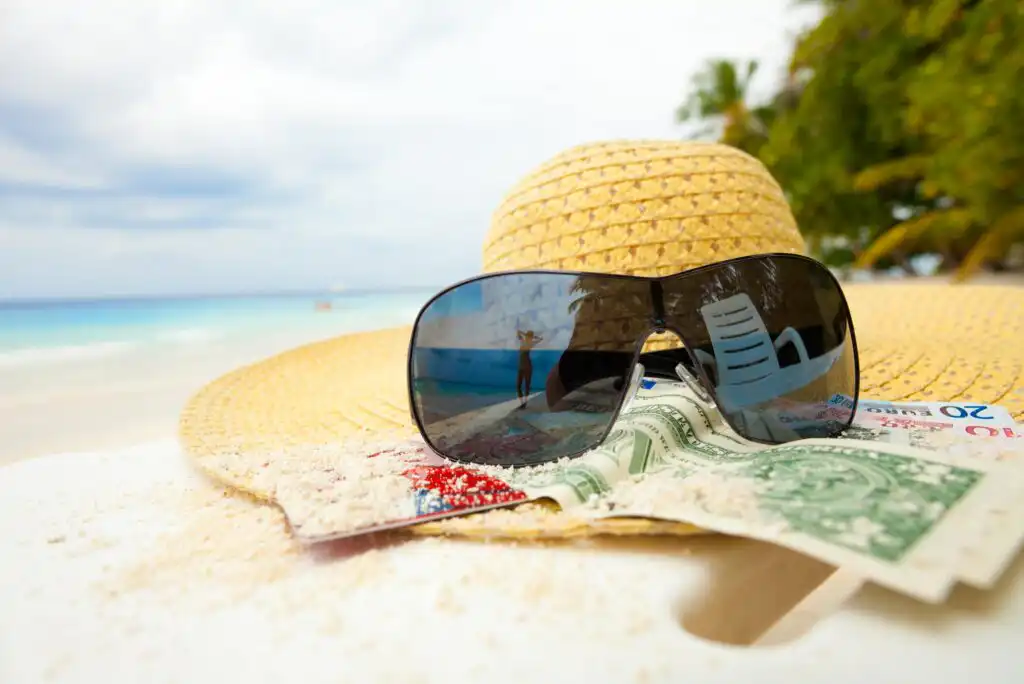 A straw hat, sunglasses, and money rest on a sandy surface at the beach, with the sea and palm trees blurred in the background. Reflections in the sunglasses show part of a lounge chair and the ocean.