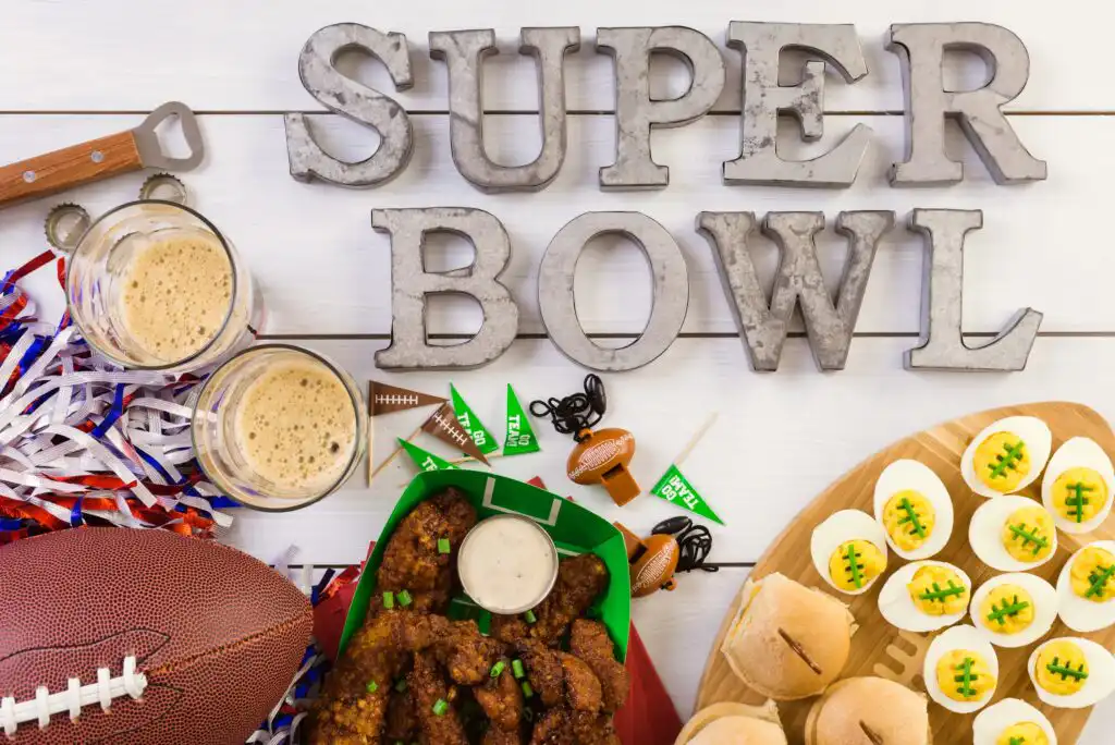 Super Bowl party setup with beer, football, chicken wings, sliders, deviled eggs decorated like footballs, and festive decorations on a white table. The words SUPER BOWL are displayed at the top.