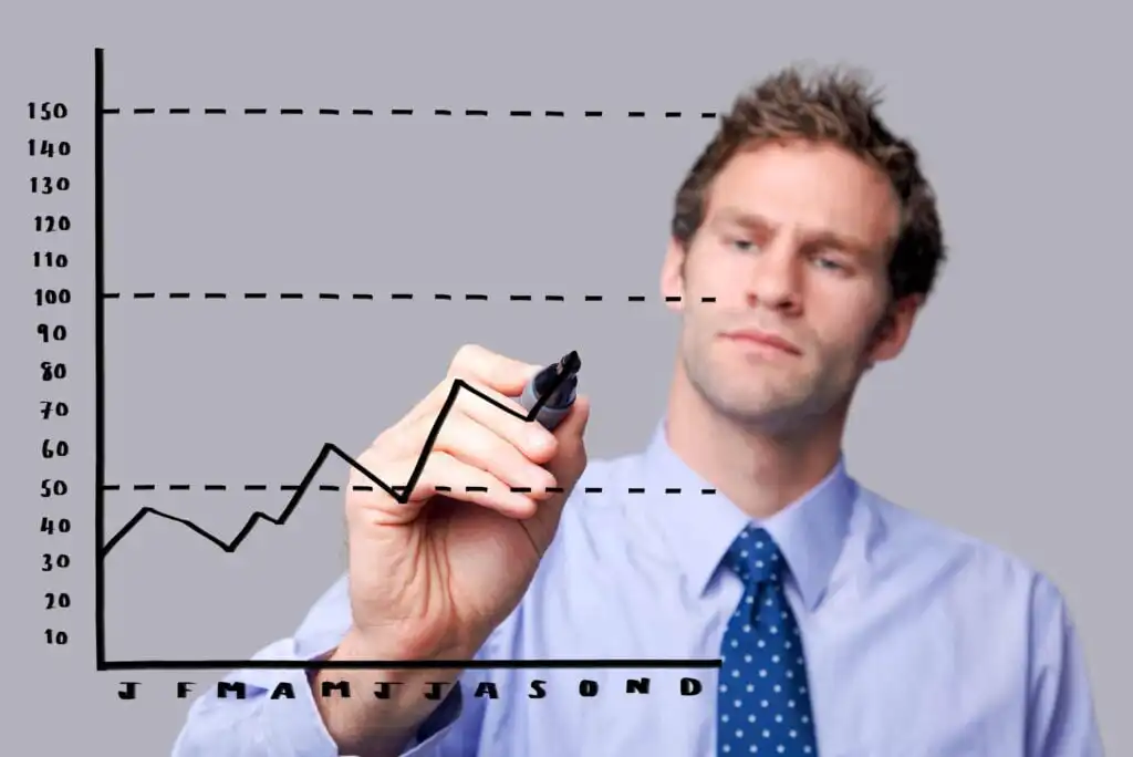 A man in a blue shirt and tie draws a line graph on a transparent screen, with months labeled along the bottom and numerical values on the side. The graph shows fluctuating data.