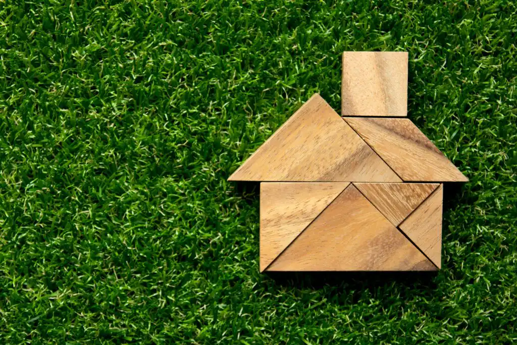 Wooden tangram puzzle pieces arranged to form the shape of a house, placed on a background of green grass.