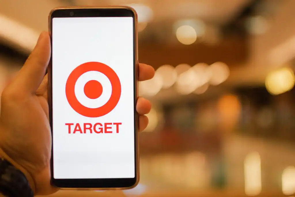 A hand holds a smartphone displaying the Target logo, which features a red bullseye above the word TARGET, against a blurred indoor background with warm lighting.