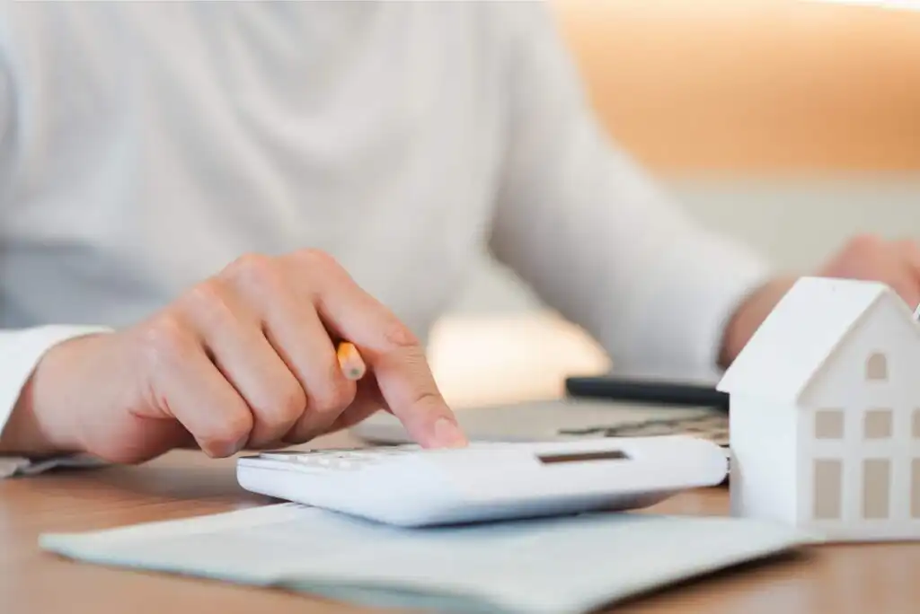 Close-up of a person’s hand using a calculator with a pencil, next to paperwork and a small white model house, suggesting financial planning or budgeting for a home.