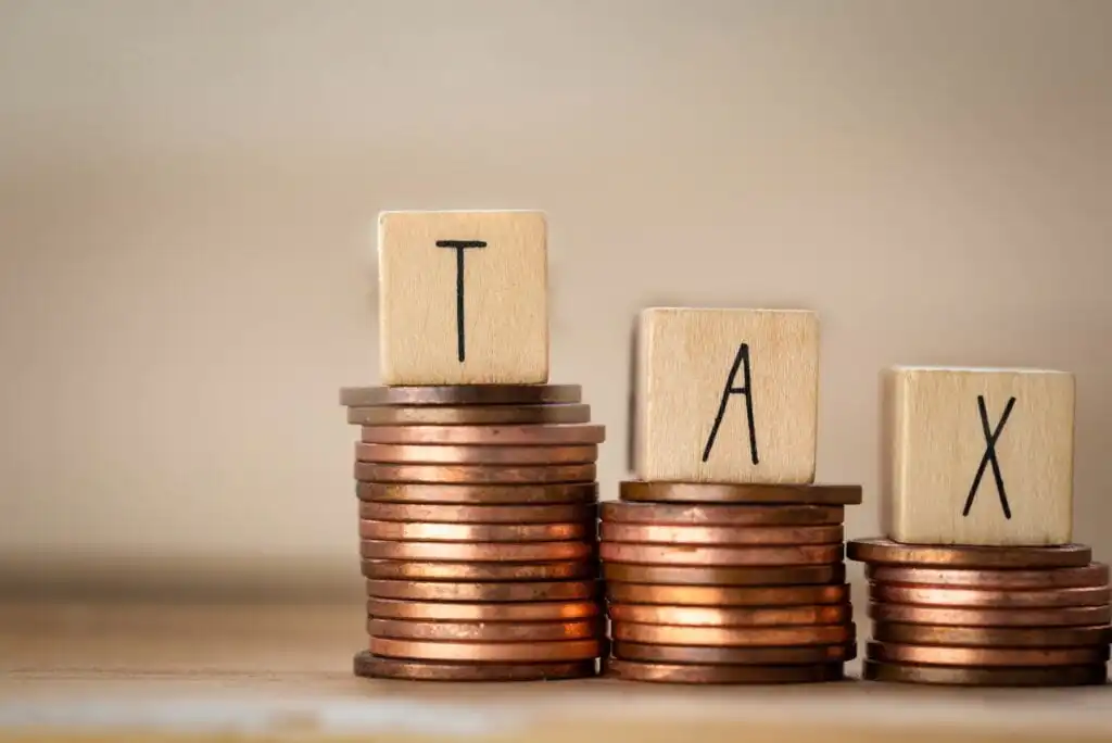 Stacks of coins with wooden blocks on top, each block displaying one letter to spell the word TAX. The background is plain and softly blurred.