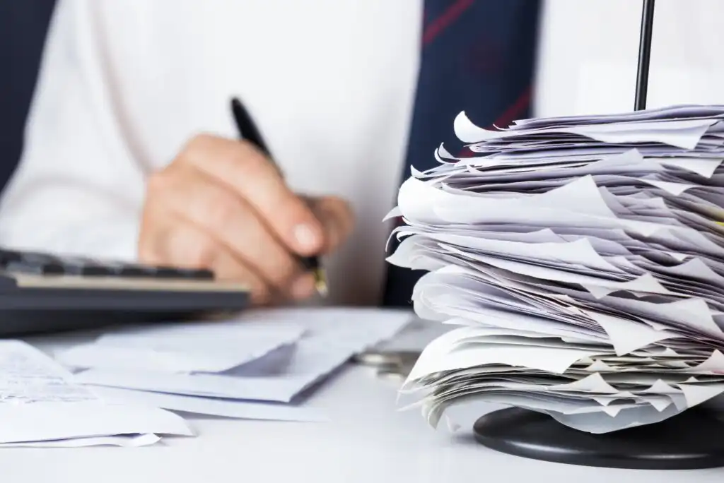 A large stack of paperwork sits on a desk in the foreground while a person in business attire writes with a pen in the background, partially out of focus.