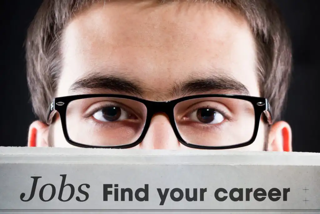 A person wearing black glasses looks over the top edge of a newspaper with the headline Jobs Find your career printed on it. The background is dark and the focus is on the persons eyes and the text.