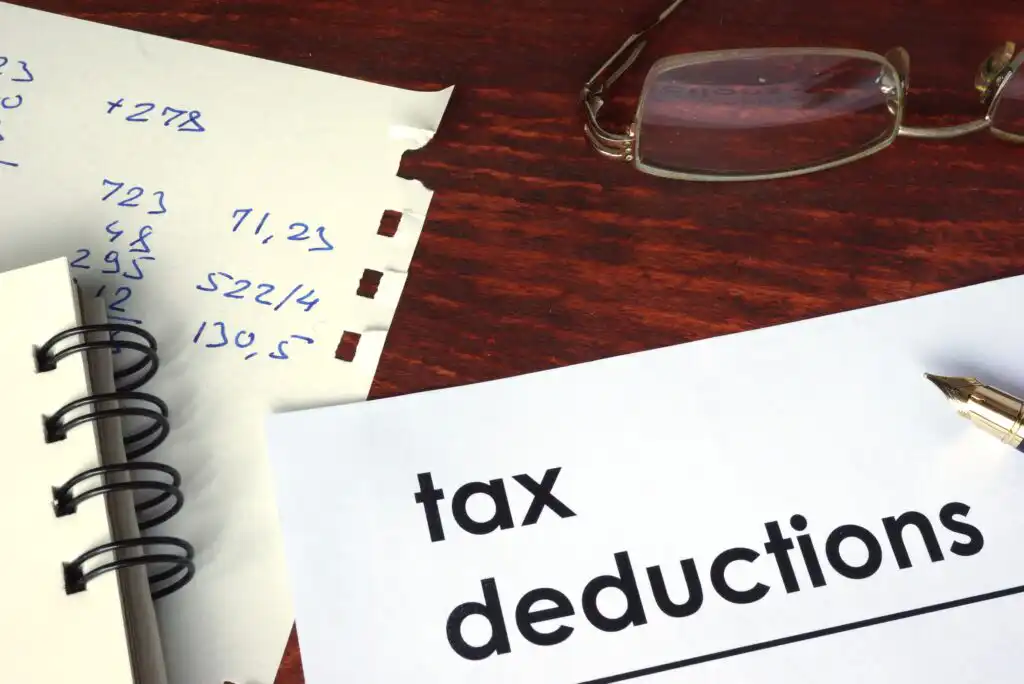 A paper labeled tax deductions lies on a wooden desk next to a pen, eyeglasses, a spiral notebook, and a torn sheet with handwritten numerical calculations.