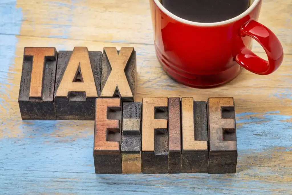 Wooden letter blocks spell out TAX E-FILE on a blue and tan wooden surface, next to the edge of a red coffee cup filled with black coffee.