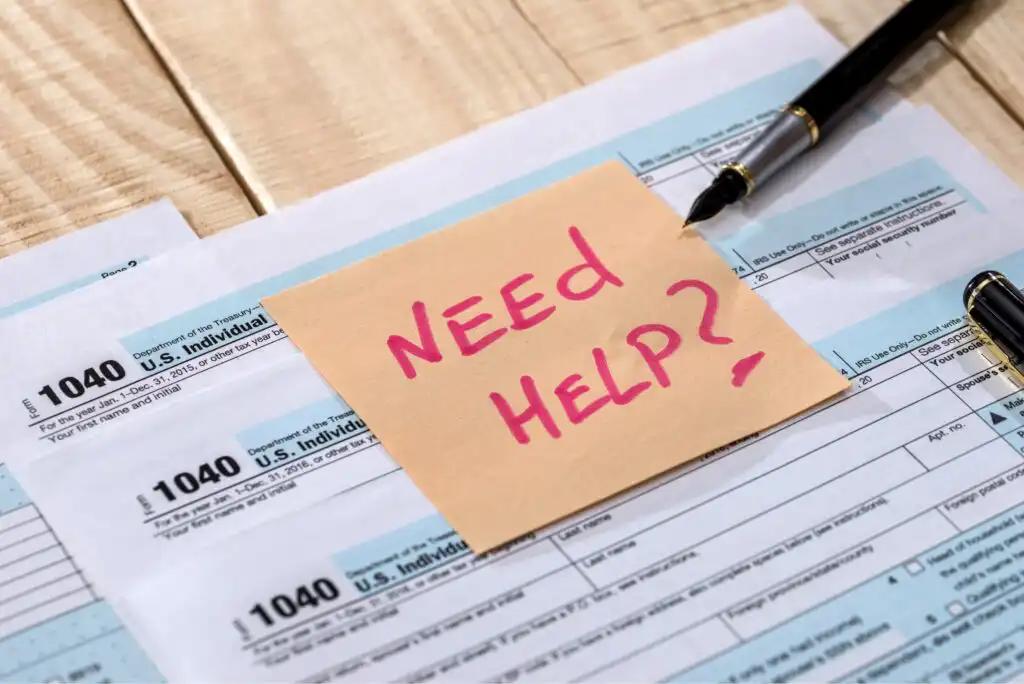 A sticky note with NEED HELP? written in pink marker lies on top of U.S. 1040 tax forms, with a black pen nearby on a wooden table.