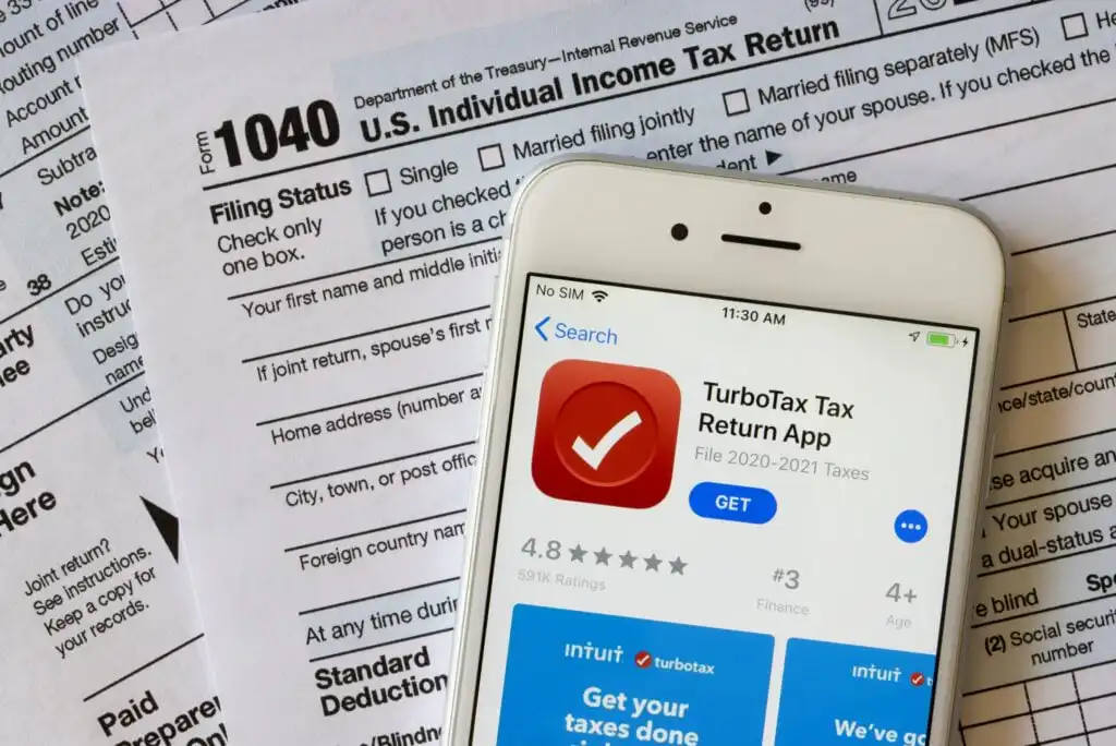 A smartphone displaying the TurboTax Tax Return App download page sits on top of U.S. 1040 Individual Income Tax Return forms.