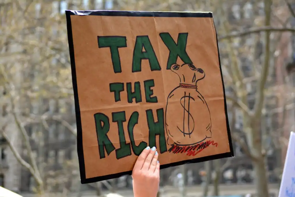 A hand holds a protest sign reading TAX THE RICH in bold letters, next to a drawing of a money bag with a dollar sign, against a blurred outdoor background.