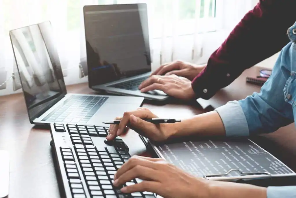 Two people working at a desk with laptops and a desktop computer, typing on keyboards and reviewing code on paper, suggesting teamwork in a programming or software development environment.