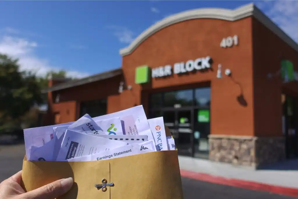 A hand holding an envelope filled with tax and financial documents in front of an H&R Block office building.