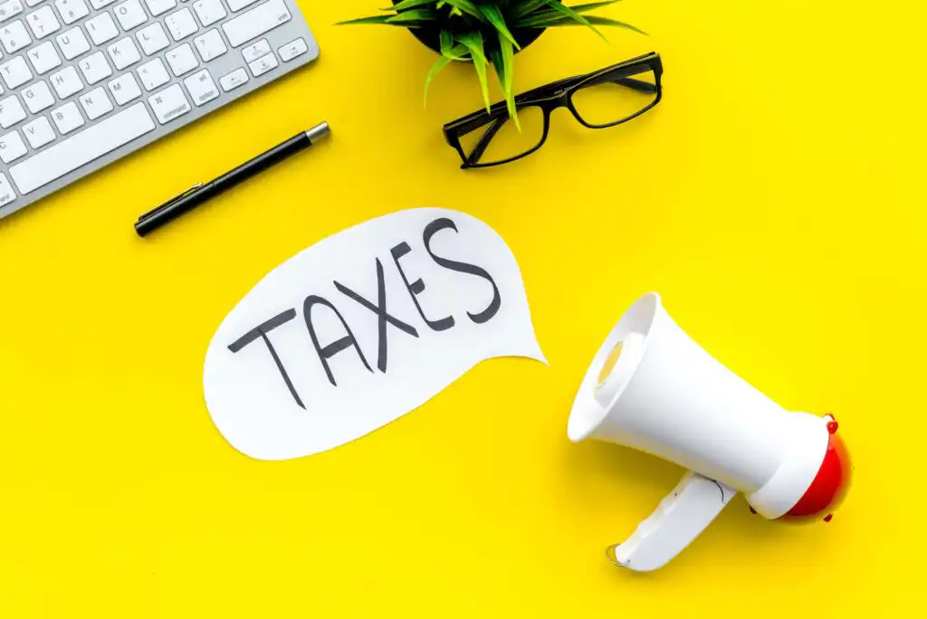 A speech bubble with the word TAXES is next to a megaphone on a bright yellow desk, alongside a keyboard, black glasses, a pen, and a small green plant.