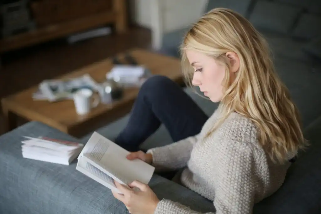 A young woman with blonde hair sits on a couch, reading a book. She is wearing a light-colored sweater and jeans. An open book and some papers are on the coffee table in the background.