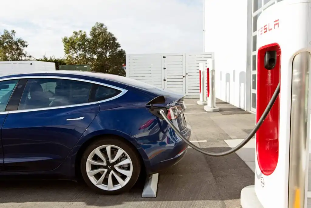 A blue Tesla electric car is parked at a charging station, plugged in and charging outdoors on a sunny day, with white buildings and trees in the background.
