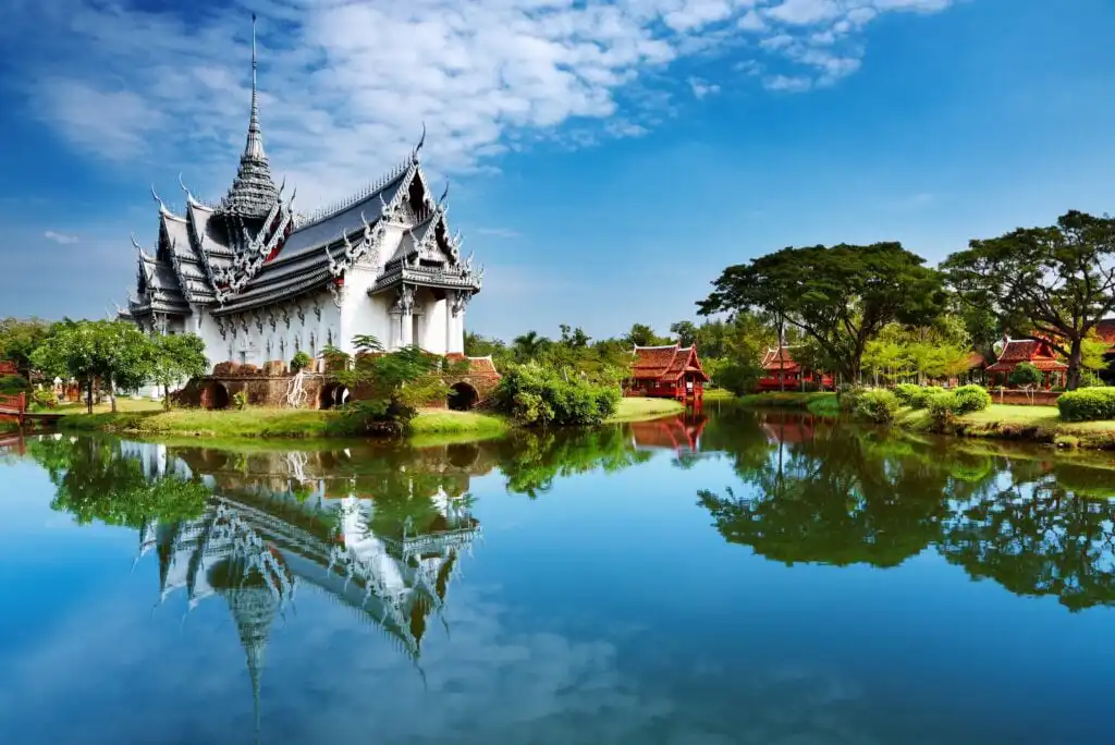A traditional Thai temple with ornate rooftops stands beside a calm pond, reflecting the building and blue sky. Lush trees and smaller pavilions surround the temple, creating a serene, picturesque scene.
