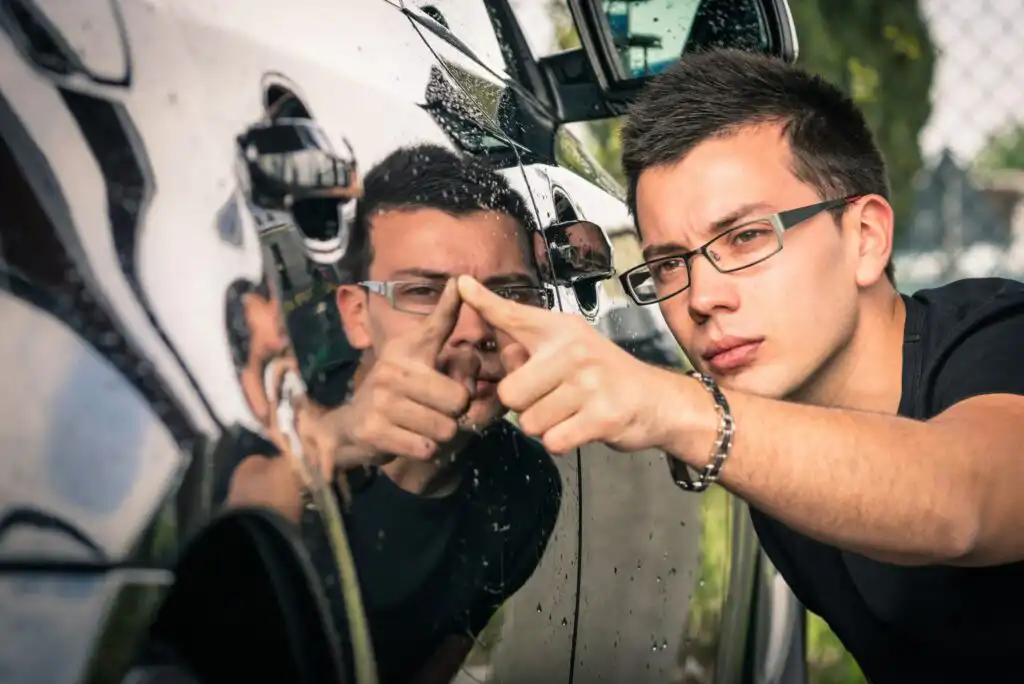 A man wearing glasses inspects the surface of a black car, closely examining a dent or blemish with his finger while his reflection appears in the car’s shiny exterior.