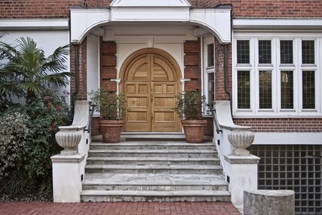 A large wooden double door with an arched top is framed by brick walls and white trim. Stone steps, flanked by potted plants and decorative urns, lead up to the entrance of a classic brick building.