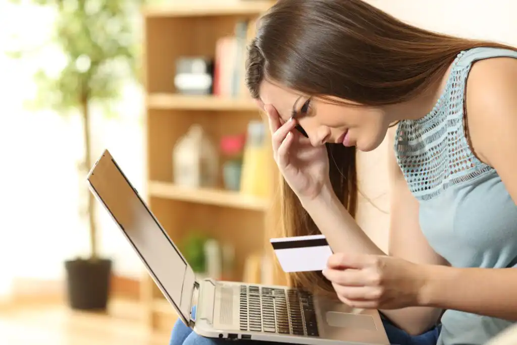 A woman sits in front of a laptop, holding her head with one hand and a credit card in the other, looking worried or stressed, with shelves and a plant in the background.