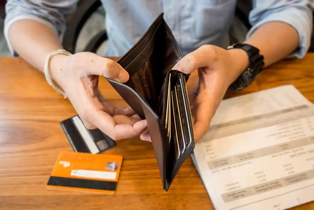 A person in a blue shirt holds an open, mostly empty wallet over a wooden table with credit cards and a bill or receipt laying on the table.